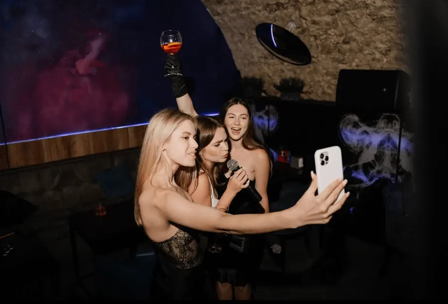 Three friends taking a selfie in the lounge, one holding a microphone and one raising a cocktail