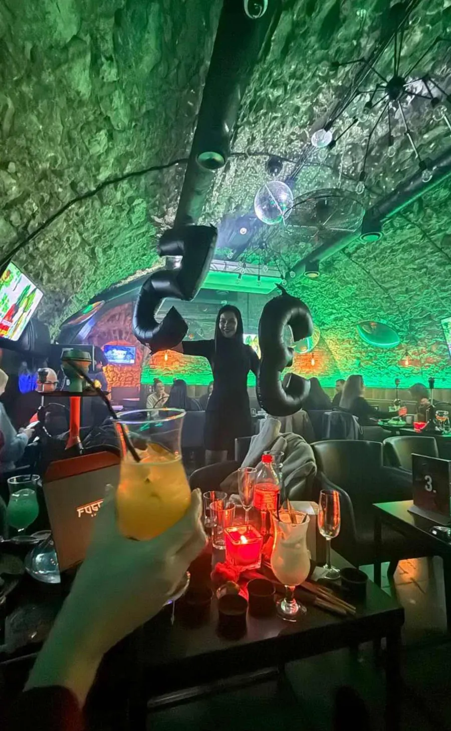 Festive celebration with silver balloons, a raised cocktail and guests under green party lighting